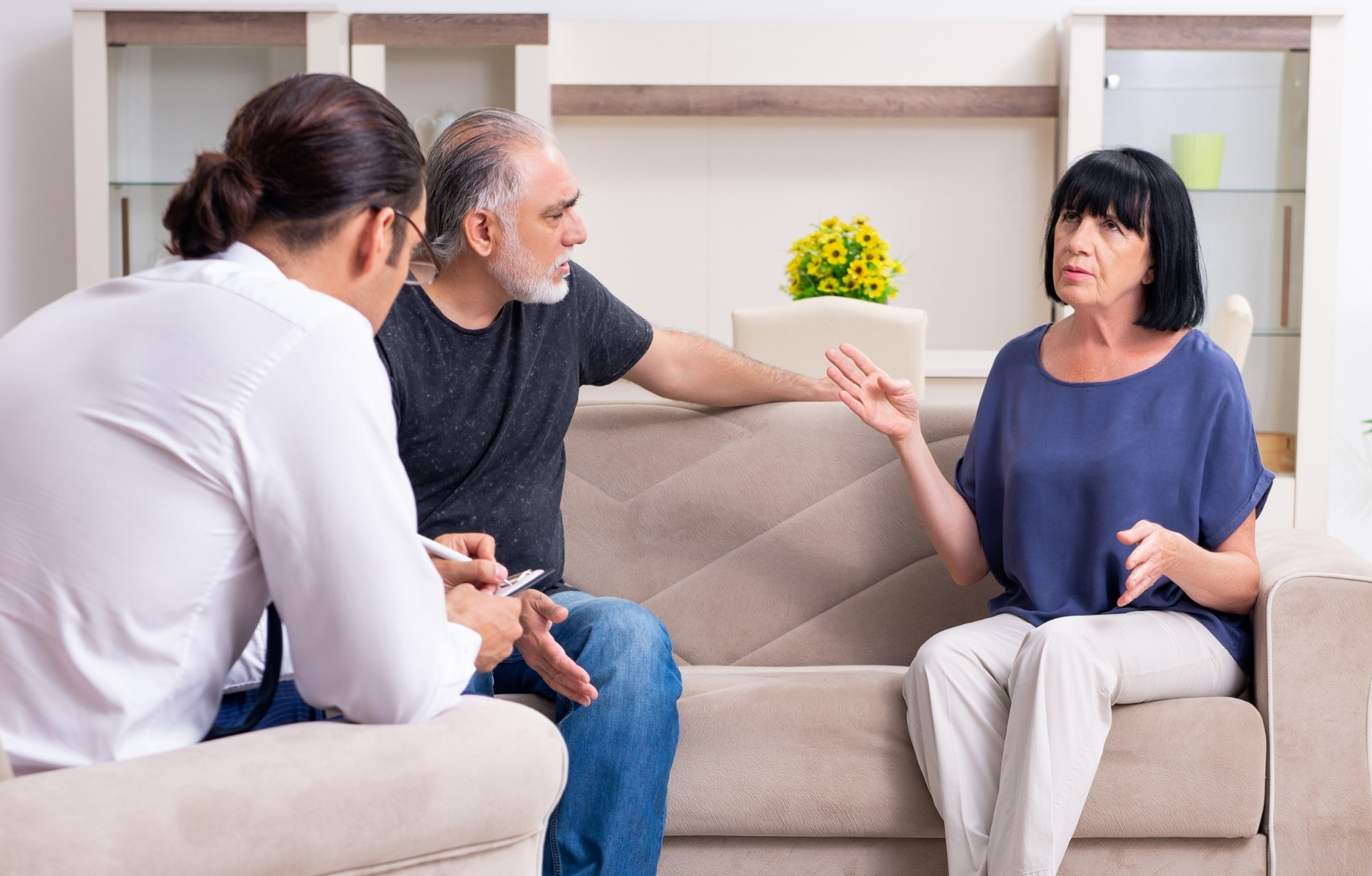 Zwei Personen sitzen auf einem Sofa, eine davon erzählend. Eine dritte Person sitzt ihnen gegenüber.